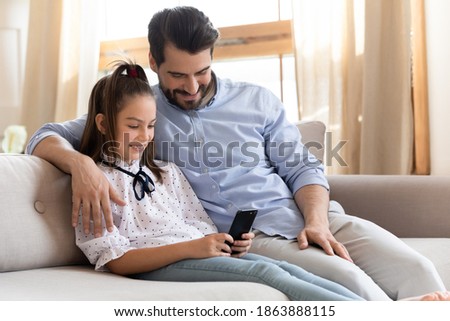 Loving Caucasian dad and small daughter sit rest on sofa in new cozy home hug enjoy family weekend time using cellphone. Happy caring father and girl child relax on couch browse modern smartphone.