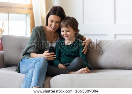 Loving young Caucasian mom and small son relax on couch in living room watch video on smartphone together. Happy caring mother or nanny and little boy child rest on sofa using modern cellphone.
