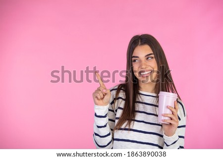 Smiling woman poses with disposable cup of hot beverage, enjoys aromatic drink in morning before going out,  pointing with finger at blank space for advertising, isolated on pink studio background