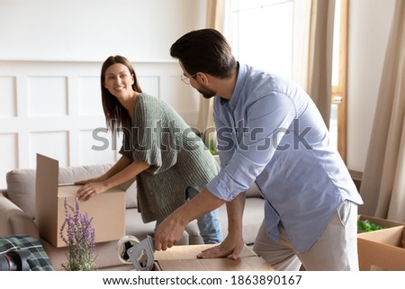 Happy young couple renters packing on moving day wrap packages parcels with adhesive scotch in living room. Excited Caucasian family tenants relocate seal boxes with tape. Delivery concept. Royalty-Free Stock Photo #1863890167