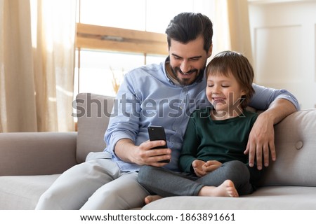 Happy caring young Caucasian father sit rest on sofa at home with excited little son watch funny video on cellphone together. Smiling loving dad relax on couch with boy child using smartphone gadget.