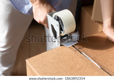 Crop close up of husband pack box with tape dispenser relocating to new apartment house. Man renter or tenant wrap package with personal belongings with adhesive scotch, moving. Delivery concept. Royalty-Free Stock Photo #1863892354