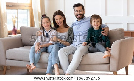 Portrait of smiling young Caucasian family renters with small kids sit rest on comfortable couch in new renovated deign home. Happy parents tenants with children relax on sofa. Rent, realty concept.