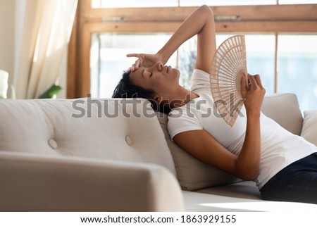 Tired unwell young African American woman sit lie on couch at home use waver suffer from hot weather indoors. Overheated millennial biracial female renter breathe fresh air from hand fan. Royalty-Free Stock Photo #1863929155