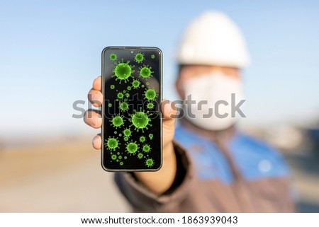 Worker is holding a mobile phone with coronavirus (covid-19) icons in the construction site. Covid-19 spreads via a number of means, primarily involving saliva and other bodily fluids and excretions.