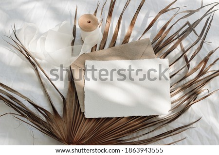 Tropical stationery still life. Closeup of blank card mock-up and craft envelope in sunlight. Dry palm leaf , white linen table cloth background. Summer vacation concept. Boho design. Flat lay, top.