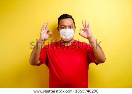 Satisfied young man in medical mask showing make okay sign against yellow background