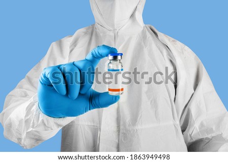 Photo of a doctor in protective suit, mask and gloves holding a vaccine bottle on blue background.