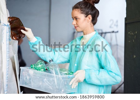 Careful attentive woman worker in protective gown sorting plastic bottles at the waste sorting plant. Stock photo. Recycling and waste sorting concept