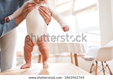 Caucasian baby taking first steps walk forward on the table. Happy little baby learning to walk with mother help at home. Mother teaching how to walk gently. Baby growth and development concept