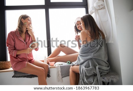 Portrait of smiling girl with mirror in hands using jade roller for skin treatment while laughing with her best friends. Stock photo