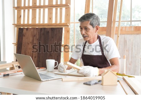 Portrait Senior asian elderly old man wearing white gloves using computer laptop to research and planning woodwork in carpentry workshop