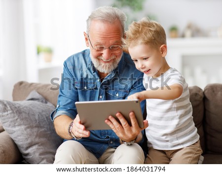 Happy family senior grandfather and boy sitting on couch and watching cartoon on tablet on weekend day at home together
