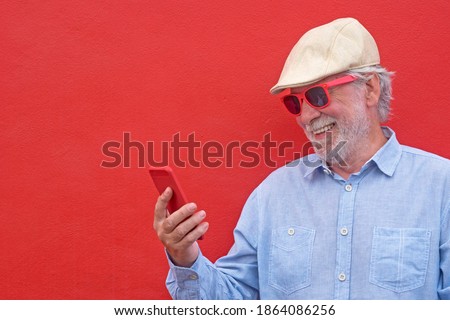 Portrait of senior white haired man with cap smiling using his smart phone, standing against red background
