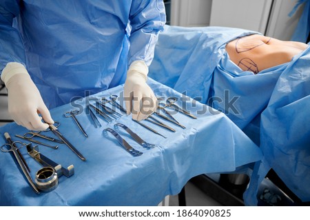 Close up of surgeon in sterile gloves getting ready medical instruments. Female patient with marks on skin lying on medical bed while doctor preparing tools. Concept of plastic surgery preparation. Royalty-Free Stock Photo #1864090825