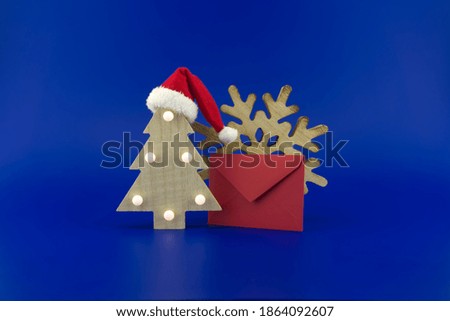 Christmas tree with a Santa hat on top next to a red envelope for greetings and a snowflake decoration on a festive blue background. New Year and Christmas greeting season concept