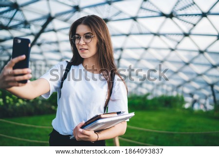 Attractive female student with education equipment using from mobile camera for clicking selfie images during pastime in orangery, beautiful girl in glasses posing for photographing herself
