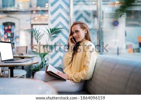 Side view of happy attractive female smiling and speaking on smartphone with notebook on knees sitting at table with laptop at cafe in Barcelona city looking at camera