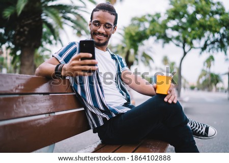 Low angle of thoughtful male in casual clothes and glasses with cup of hot coffee sitting on bench and browsing mobile phone