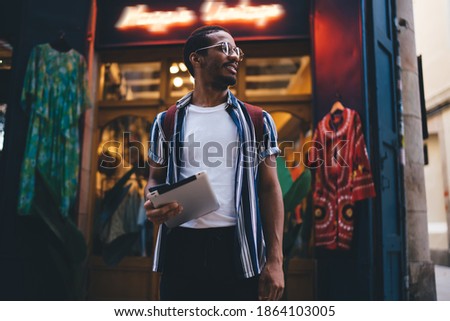 Positive trendy dressed african american male standing on street with retro neon banner gosling digital tablet, smiling dark skinned hipster guy explore destination using touchpad during journey
