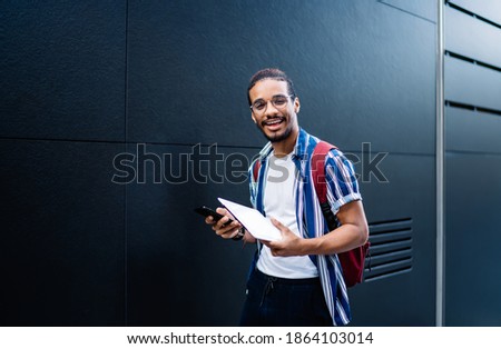 Cheerful trendy male student wearing casual outfit and eyeglasses walking down city street using tablet and chatting via smartphone smiling at camera