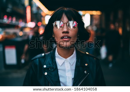 Caucasian woman in optical spectacles with neon reflection of lights standing at urbanity during travel vacations for visiting New York, attractive hipster girl in trendy apparel going out Royalty-Free Stock Photo #1864104058