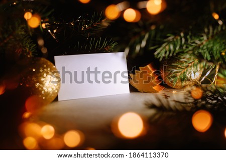 Blank business card with christmas tree branches and golden garland lights. New year celebration concept banner. Gift certificate. Side view. Cozy frame decoration. Invitation to an event or party. Royalty-Free Stock Photo #1864113370