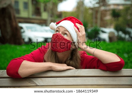 Caucasian blond girl in red hat, coat and red protective face mask sitting outdoors. Thinking of Christmas 2020. Celebrating new year in pandemic world. fashion mask for celebration. green grass in