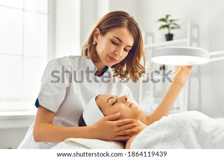 Young woman cosmetologist or dermatologist directing lamp for facial treatment to lying young woman in beauty spa salon. Facial treatment, massage, skincare, cosmetology concept Royalty-Free Stock Photo #1864119439