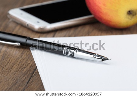 fountain ink pen with cap on table with sheets of white paper and phone with red apple