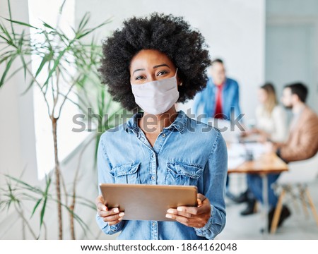 portrait of a smiling young businesswoman wearing a mask and holding a tablet in the office Royalty-Free Stock Photo #1864162048