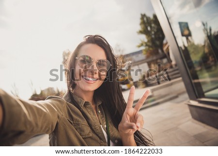 Photo of adorable sweet young lady wear brown shirt dark glasses smiling making selfie showing v-sign outside sunny street