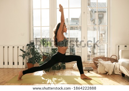 Side view of mindful barefoot young female in active wear standing on yoga mat in warrior one pose, doing Surya Namaskar or Sun Salutation in the morning. Energy, strength, balance and wellness Royalty-Free Stock Photo #1864222888