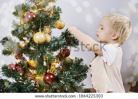 A small boy with white hair stands at the elegant Christmas tree and looks at toys. Ornaments on Christmas tree with light, bauble