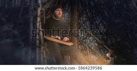 A bearded man -a tramp, a forester or a hermit, built a hut out of branches with an axe in the forest, lit a fire, and plays kantele (gusli, psaltery) at night. Wide cinematic image. Royalty-Free Stock Photo #1864239586