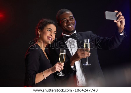 Waist up portrait of elegant mixed-race couple taking selfie photo while standing against black background at party, copy space