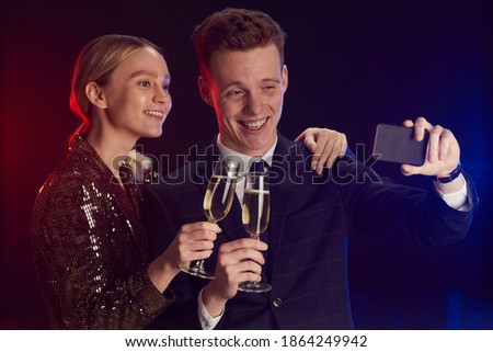 Waist up portrait of young couple taking selfie photo via smartphone while enjoying party at prom night standing against lack background
