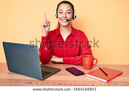 Young brunette woman wearing operator headset at the call center office smiling happy pointing with hand and finger to the side 