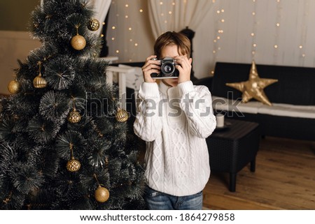 A cute child photographs you on a retro camera against the background of an artificial Christmas tree with golden balls. A little boy is playing and having fun with a camera