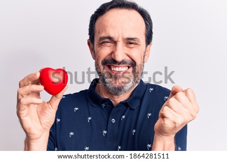 Middle age handsome romantic man holding plastic heart over isolated white background screaming proud, celebrating victory and success very excited with raised arm