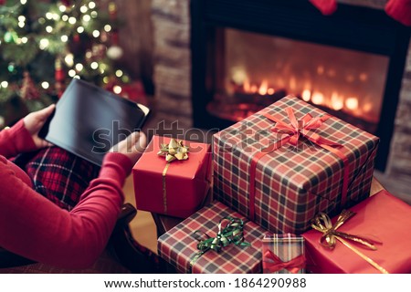 Christmas. Woman in sweater using tablet for searching gift ideas sitting at table near fireplace and christmas tree. Concept
