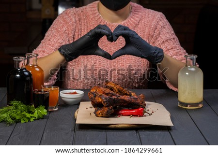 Young girl in pink sweater and black gloves shows with his hands a shape in the form of a heart before eating fried ribs with pepper and bottle of wine