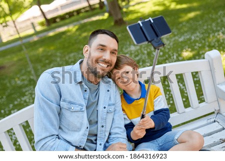 family, fatherhood and people concept - happy father and his little son taking picture with smartphone and selfie stick sitting on bench at summer park