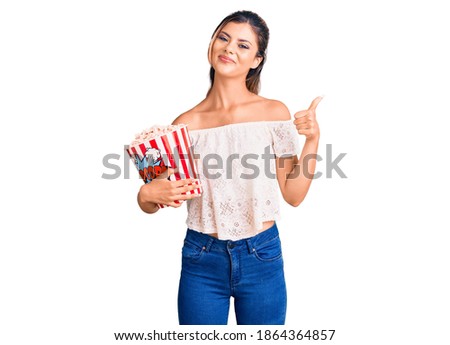 Young beautiful woman holding popcorn smiling happy and positive, thumb up doing excellent and approval sign 