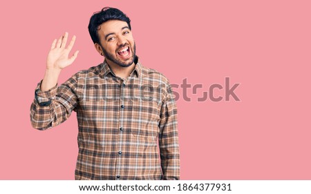 Young hispanic man wearing casual clothes waiving saying hello happy and smiling, friendly welcome gesture 