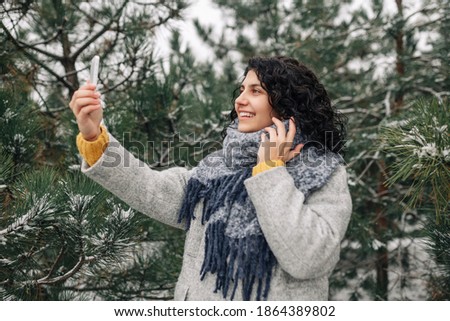 Smiling young woman taking selfie at the snowy winter park. Female taking a photo of herself with a mobile phone wearing a grey coat and blue scarf among fir trees cover with snow. Happiness concept.