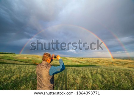Elderly man taking pictures on a smartphone he is a magnificent rainbow Above the steppe Spring day.