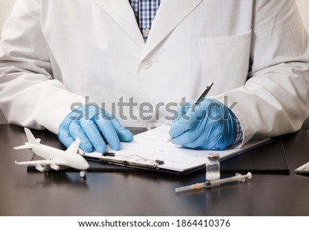 Doctor's hand with pen filling in data, on form, with white plane, syringe,, computer keyboard, on work table, white background.