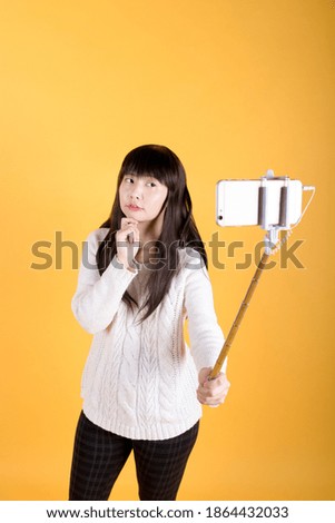Woman blogger holding up her phone on a selfie stick to take picture or video of herself in studio on yellow background.