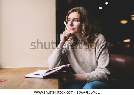 Young female novelist in baggy sweater thinking hand at chin and looking away through window while sitting at cafe table with open diary and pen in hand Royalty-Free Stock Photo #1864435981